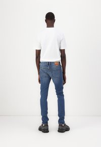 Blue denim jeans with a straight fit, featuring a leather logo patch at the back, worn with a plain white t-shirt and black sneakers.