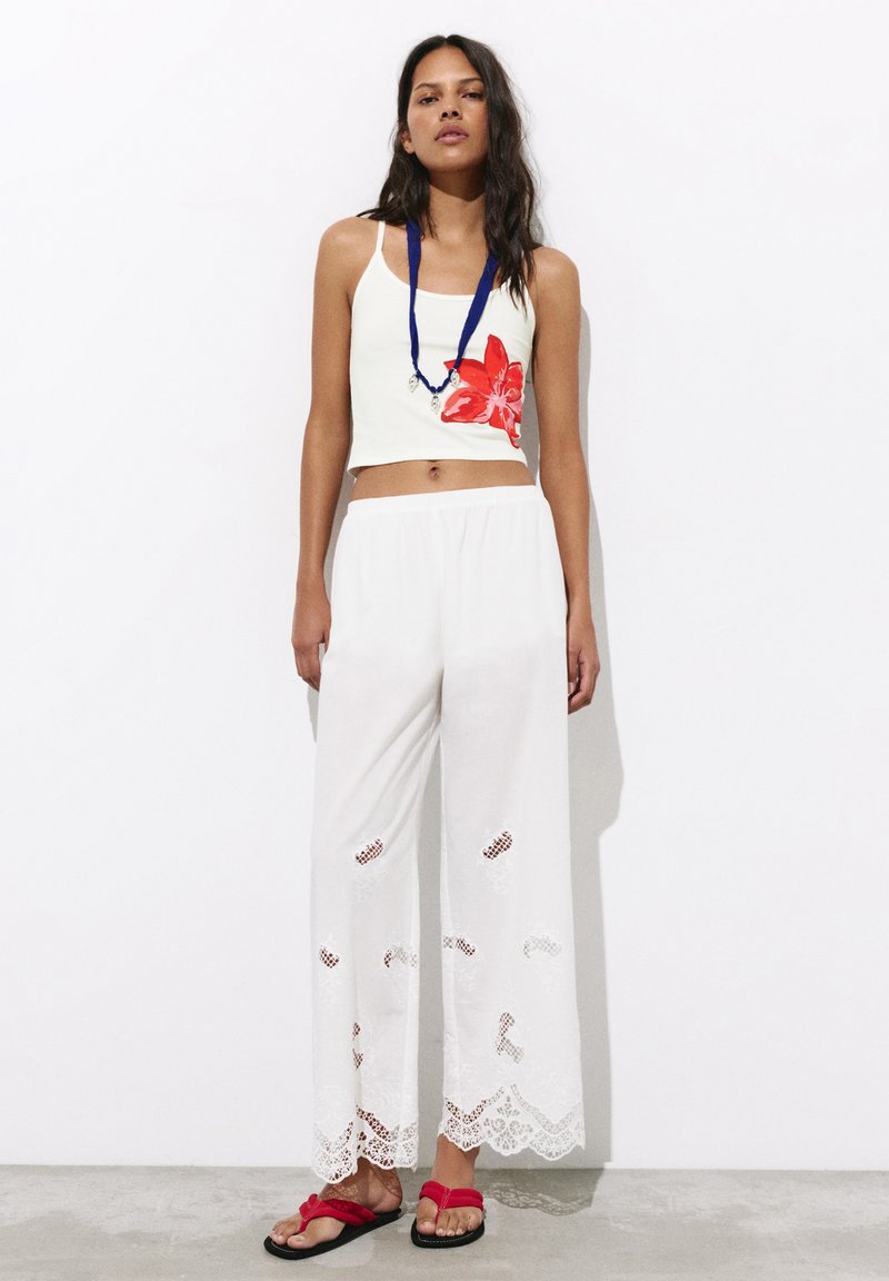 Femme portant un crop top blanc avec un imprimé fleur rouge, un pantalon blanc à jambes larges avec des découpes en dentelle, des sandales rouges et un collier bleu, debout contre un mur blanc.
