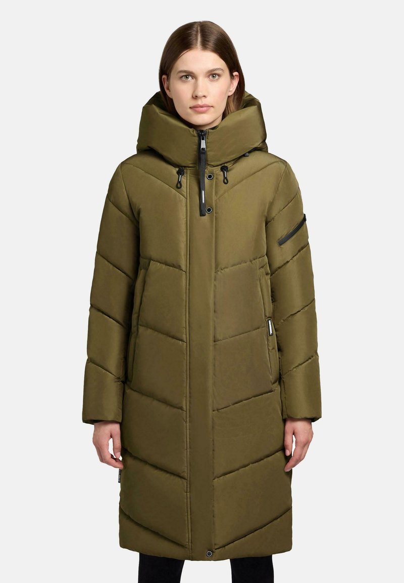 Kvinna i en olivgrön lång pufferjacka med hög krage, framdragkedja och ficka med dragkedja på vänster ärm, som står framför en vit bakgrund.