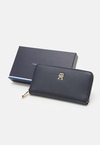 Marineblaues Lederportemonnaie mit goldfarbenem Logo, Reißverschluss und schlankem Design. Inklusive einer passenden schwarzen Box mit buntem Streifenakzent.