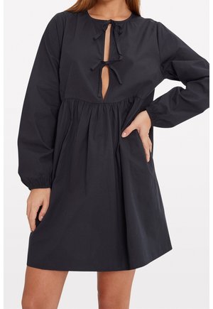 Woman wearing a loose black long-sleeve dress with two front tie closures and elastic cuffs.