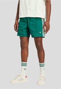 Grønne sports shorts med hvide sidelister og et guld logo. Parret med hvide sokker med grønne striber og hvide sneakers.