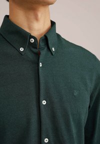 Temnozelena srajca z gumbi iz teksturirane tkanine, s koničastim ovratnikom, belimi gumbi in vezenim logotipom na prsih.