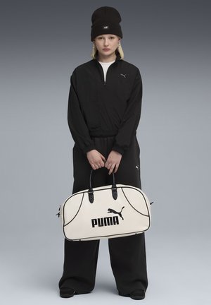Zwarte gecentraliseerde jas en broekset met elastische manchet, gecombineerd met een beige sporttas met een zwart "PUMA" logo en donkere handvatten.