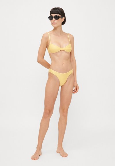 Set di bikini giallo con finitura testurizzata; top stile bralette con spalline sottili e slip alti coordinati. Indossato con occhiali da sole neri.