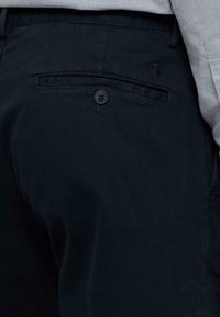 Pantaloni blu scuro realizzati in un tessuto liscio, con una tasca posteriore con chiusura a bottone e un logo distintivo sopra la tasca.