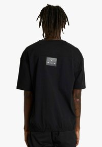 Persona con dreadlocks che indossa una maglietta nera a maniche corte e larga con una piccola toppa rettangolare sulla parte superiore della schiena e orlo elastico.