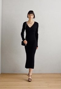 Rochie neagră tricotată cu nervuri, cu mâneci lungi și decolteu în V. Rochia ajunge până la jumătatea gambei și are o croială ajustată. Sandale negre cu bretele.