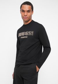 Melns garas piedurknes T-krekls ar "GUESS" logo metāla fontā. Mīksta auduma, ar apaļu kakla izgriezumu un brīvu piegriezumu. Stilizēts ar tumšām biksēm.
