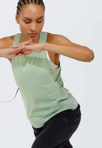Canotta sportiva verde chiaro con scollatura rotonda e spalline sottili, indossata sopra una canotta bianca, abbinata a pantaloni neri. Nessun motivo visibile.