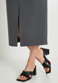 Jupe grise longueur genou avec une fente latérale, réalisée en tissu texturé. Associée à des sandales à talons blocs noires avec des brides croisées.