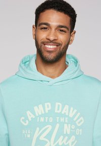 Sudadera azul claro con cordón y gráfico frontal: "CAMP DAVID INTO THE NO. 01 Blue." Textura suave y diseño casual.
