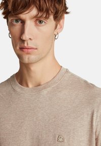 Světlé hnědé tričko s krátkým rukávem, kulatým výstřihem a texturovanou látkou. Na levé straně hrudi má malé vyšívané logo.