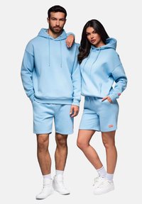 Lichtblauwe hoodie en korte broek set van zacht materiaal. Heeft een ontspannen pasvorm, verstelbare capuchon en kleine logo-accenten op de korte broek.