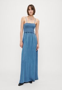 Šviesiai mėlynas džinsinis maksi suknelė su smulkintu bodice, plonomis petnešomis ir laisvai krentančia sijonu. Lygus tekstūroje su minimaliu dekoravimu.
