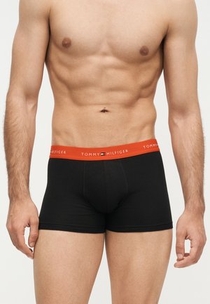 Torso masculino sin camiseta con calzoncillos bóxer negros Tommy Hilfiger con una cintura roja, de pie frente a un fondo liso.