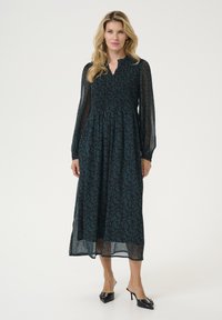 Long, teal dress with floral pattern, smocked bodice, and sheer long sleeves. Worn with black heeled mules, showcasing an elegant silhouette.
