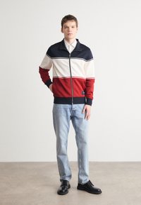 Giacca zip con strisce blu navy, crema e rosso; polsini e orlo a coste; abbinata a jeans azzurri chiari e scarpe nere; texture liscia.