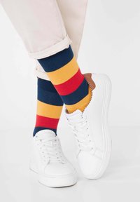 Weiße Sneakers mit glatter Oberfläche, kombiniert mit mehrfarbigen gestreiften Socken in Marineblau, Rot und Gelb. Die Hosen sind hell gefärbt und haben einen Umschlag.