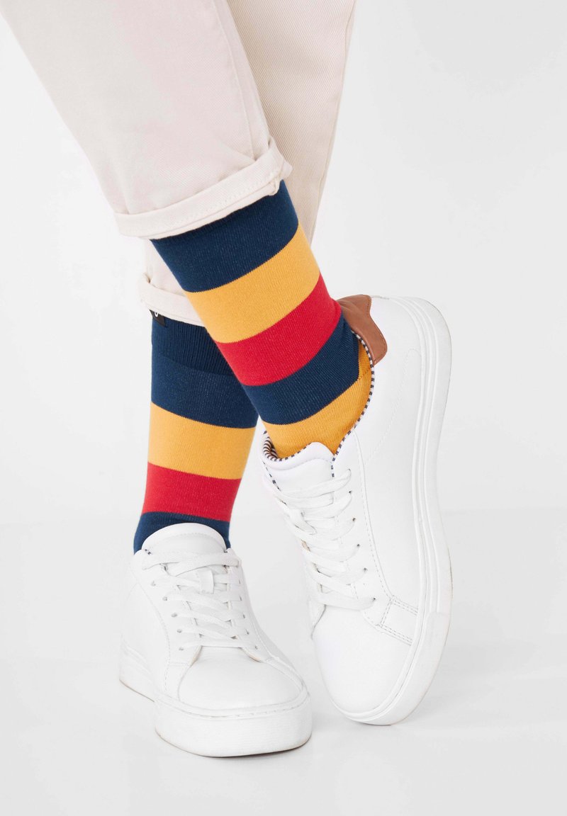 Weiße Sneakers mit glatter Oberfläche, kombiniert mit mehrfarbigen gestreiften Socken in Marineblau, Rot und Gelb. Die Hosen sind hell gefärbt und haben einen Umschlag.