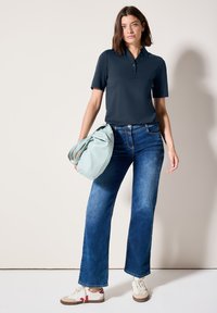 Námořnická polokošile s krátkým rukávem, spojená s modrými zvonovými džíny. Modelka drží světle zelenou tašku a má na sobě bílé tenisky s hnědými akcenty.