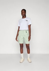 Camiseta blanca con logotipo, pantalones cortos cargo verde claro con dos bolsillos laterales, combinados con zapatillas beige y calcetines blancos. Diseño atlético simple.