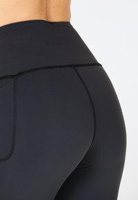 Musta korkeavyötäröinen legginsit, joissa on sileä ja joustava kangas. Varustettu litteillä saumoilla ja hillityllä logolla vyötäröllä lisäyksinä.