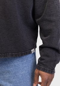 Maglietta nera a maniche lunghe con scollo rotondo, caratterizzata da cuciture a vista e un piccolo etichetta "REBEL" sull'orlo. Indossata con jeans di denim azzurro chiaro.