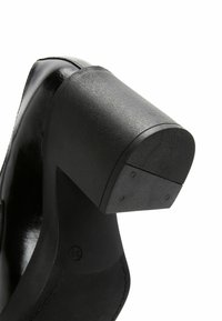 Gros plan sur une chaussure noire à talon haut, mettant en valeur le talon bloc texturé et la semelle, sur fond blanc.