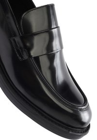 Zwarte gepolijste leren loafer met gestikte details en een brede band over de voorkant, afgebeeld vanuit een close-up schuine hoek.