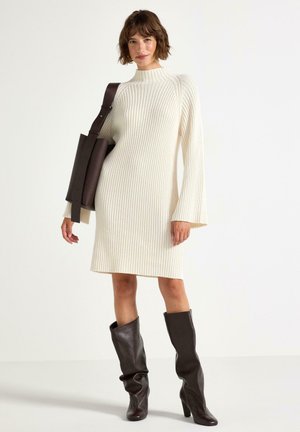 Abito maglione a coste bianco crema con colletto alto e maniche svasate. Indossato con stivali marroni fino al ginocchio e una borsa a tracolla coordinata.