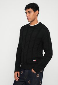 Schwarzer Strickpullover mit einem strukturierten Kabelmuster, gerippten Bündchen und Saum sowie einem kleinen Logopatch in der unteren linken Ecke.