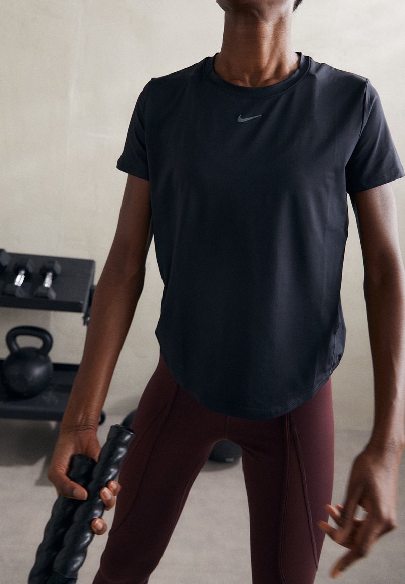 Czarny sportowy t-shirt z krótkim rękawem i zaokrąglonym dołem, w połączeniu z bordowymi legginsami. W tle widoczny sprzęt fitness.