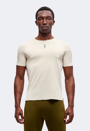Homme athlétique portant une chemise beige ajustée à manches courtes avec un logo près du col et un pantalon vert olive, debout devant un fond uni.