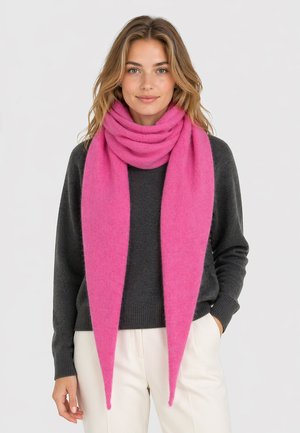 Woman wearing a long, bright pink scarf wrapped loosely around her neck, dark gray sweater, and white pants, standing against a plain background.