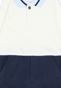 Polo à manches courtes avec un haut blanc et un bas marine, doté d'un col bleu clair et d'un bouton noir avec le numéro 8.