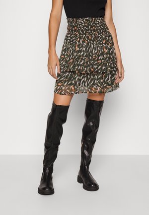 Femme portant un haut sans manches noir, une jupe à couches avec motifs vert foncé et orange, et des bottes en cuir noires montantes jusqu'aux genoux.