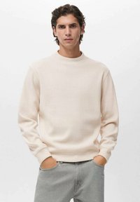 Sudadera crewneck de color beige claro hecha de un tejido de punto texturizado con mangas largas. El modelo lleva jeans grises con las manos en los bolsillos.