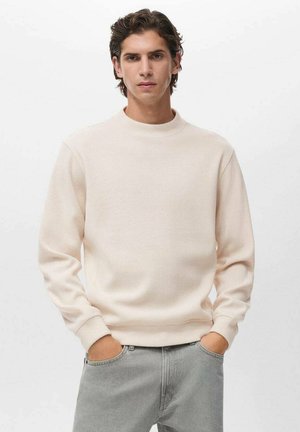 Sudadera crewneck de color beige claro hecha de un tejido de punto texturizado con mangas largas. El modelo lleva jeans grises con las manos en los bolsillos.