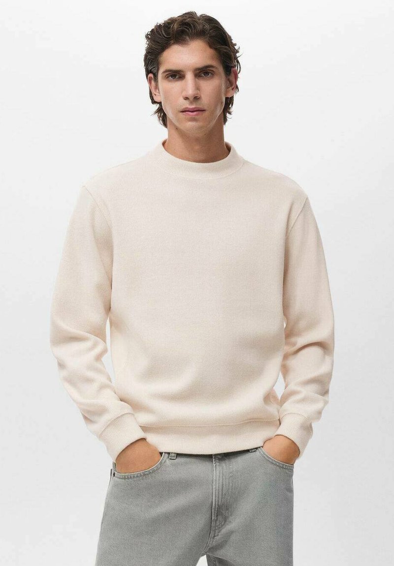 Sudadera crewneck de color beige claro hecha de un tejido de punto texturizado con mangas largas. El modelo lleva jeans grises con las manos en los bolsillos.