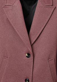 Primo piano di un blazer a spina di pesce rosa-bruno con due bottoni neri e rever a scantonatura sopra una fodera interna nera.