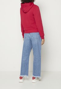 Czerwona bluza z kapturem w połączeniu z jasnoniebieskimi jeansami o szerokich nogawkach oraz białymi sneakersami z czerwonymi akcentami i postrzępionymi brzegami.