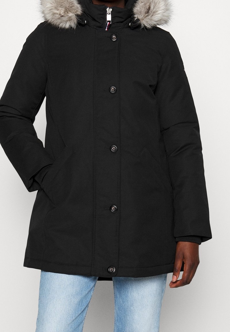 Parka d'hiver noire avec capuche bordée de fourrure, fermeture à boutons et à glissière, portée avec un jean bleu clair, main dans la poche sur un fond clair.