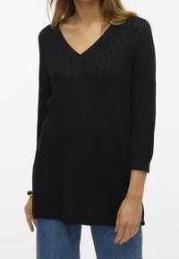 Nainen, jolla on päällään musta V-pääntie neule, jossa on kolmannes hihat, ja siniset farkut, seisoo yksinkertaista valkoista taustaa vasten.