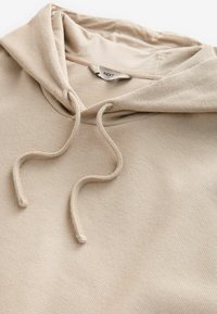 Hanorac bej cu glugă, cu o textură ribbed, sistem de închidere cu șnur și etichetă de marcă vizibilă. Material moale și design casual.