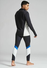 Ung man barfota klädd i svart, vitt och blått idrottstøj, står med ryggen vänd och tittar åt sidan mot en neutral bakgrund.