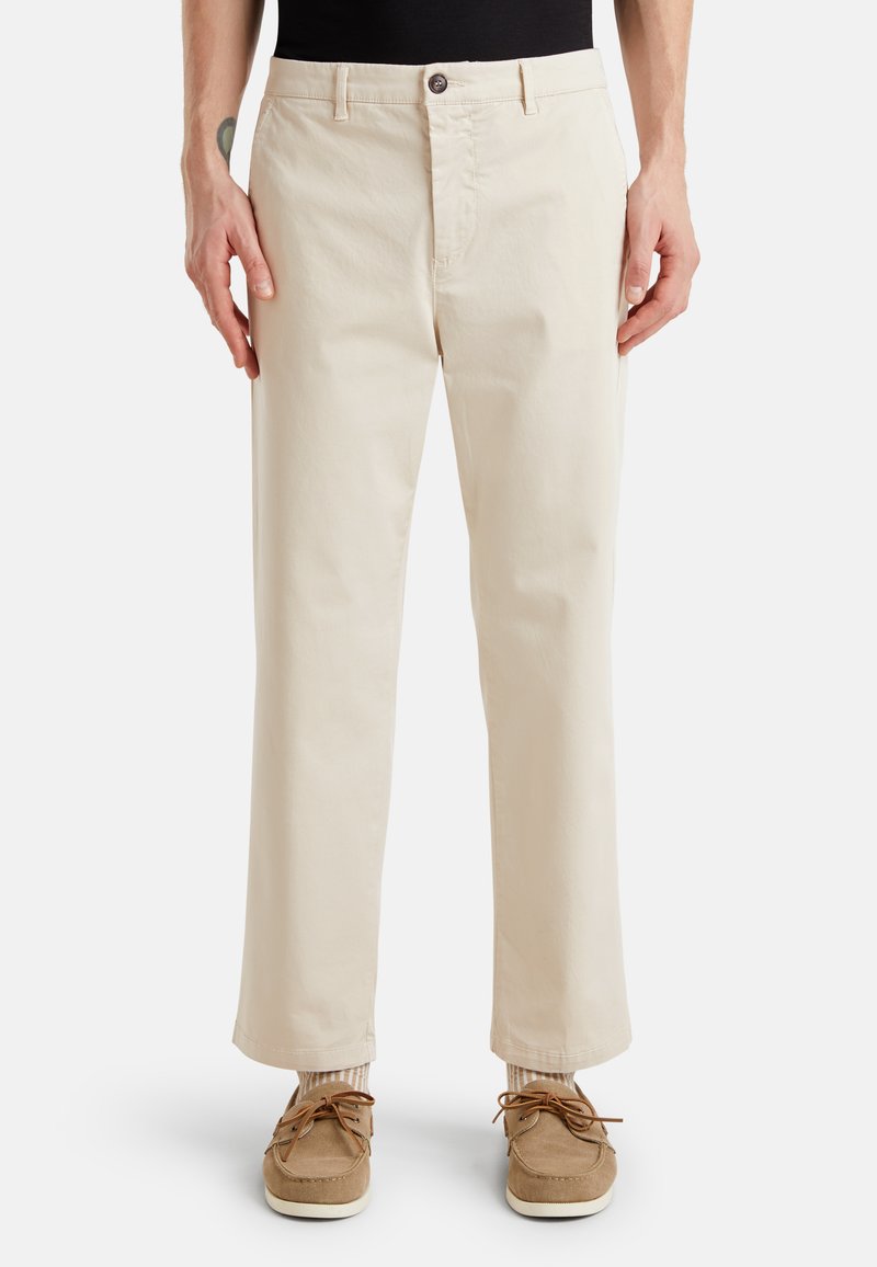 Pantalons en coton beige à coupe droite, fermeture à bouton à l'avant et poches latérales ; associés à des chaussures en suède marron et des chaussettes rayées.