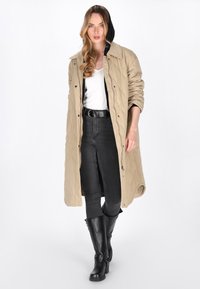 Lang beige quiltet frakke med hætte i sort, båret over en hvid top og sorte højtaljede jeans, parret med sorte knæhøje støvler.