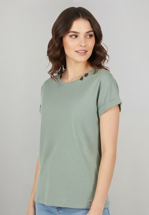 Frau mit schulterlangem braunem Haar, die ein hellgrünes Kurzarmhemd mit umgeschlagenen Ärmeln trägt und nach rechts blickt, vor einem einfarbigen Hintergrund.