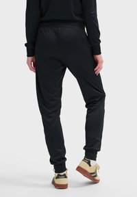 Zwarte sportbroek met een gladde textuur, elastische tailleband, omgeslagen broekspijpen en een relaxte pasvorm, gecombineerd met beige sneakers met zwarte accenten.